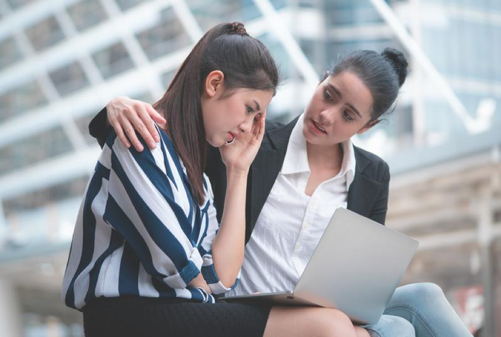 A woman is comforting another woman who is sitting in front of a laptop computer.